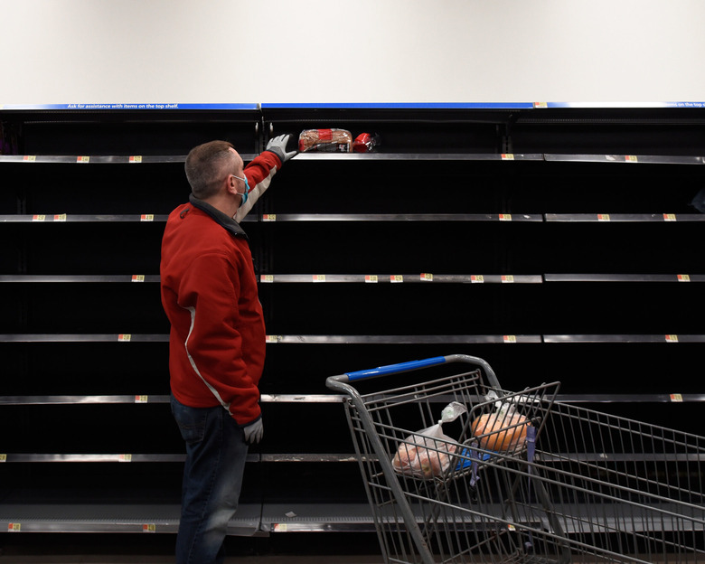 A man is wearing a medical mask at a grocery store and reaching for bread with empty shelves around him